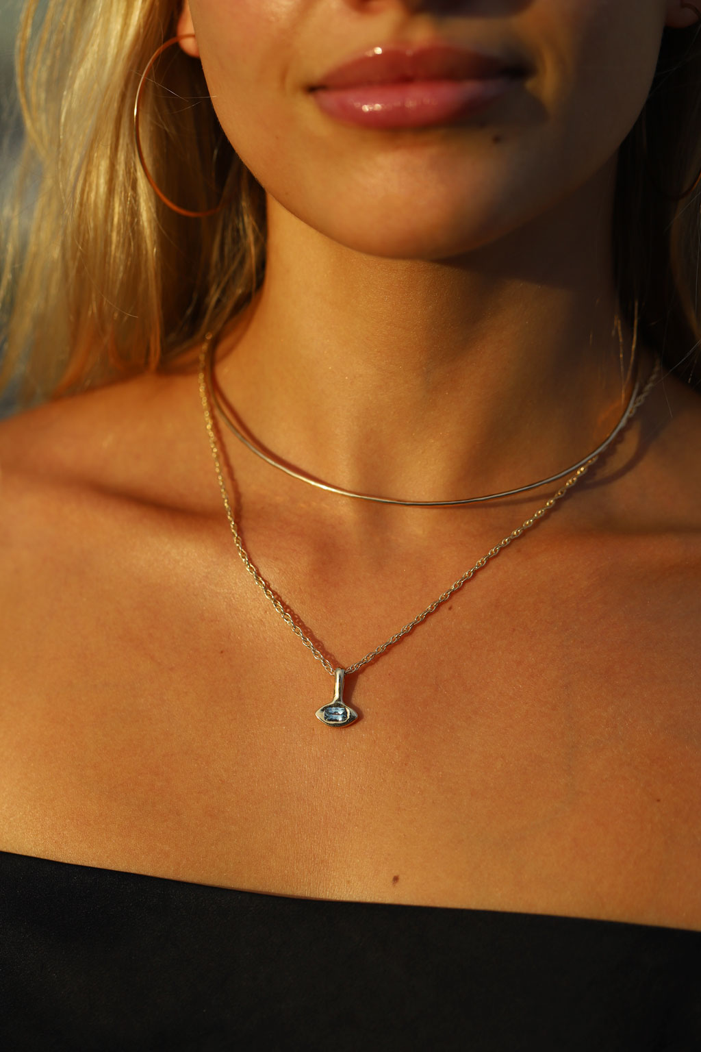 A close-up lifestyle shot of a model wearing a layered gold necklace set, featuring a delicate chain with a small silver-toned eye pendant and a thin curved chain.