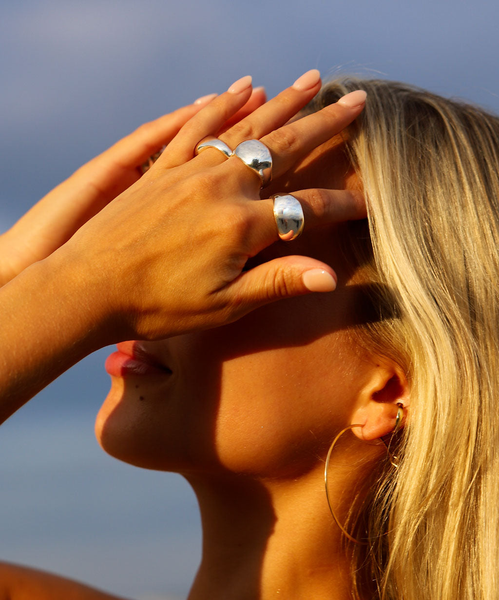 A close-up lifestyle shot of a woman outdoors at sunset, wearing several wide, polished silver rings on her fingers and a small hoop earring. Her face is partially obscured by her hands, with warm golden-hour light casting shadows against a blue sky background.