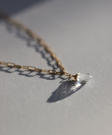 closeup side view stilllife of a crystal quartz briolette and gold paperclip chain necklace against a white paper background