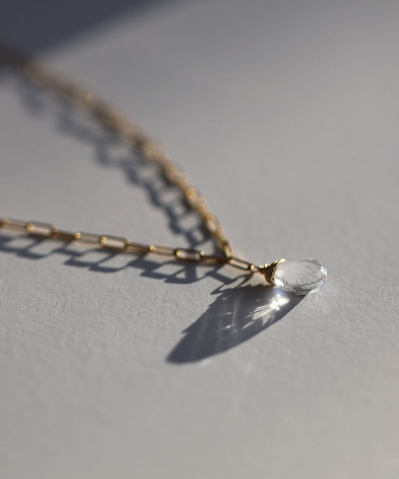 closeup side view stilllife of a crystal quartz briolette and gold paperclip chain necklace against a white paper background