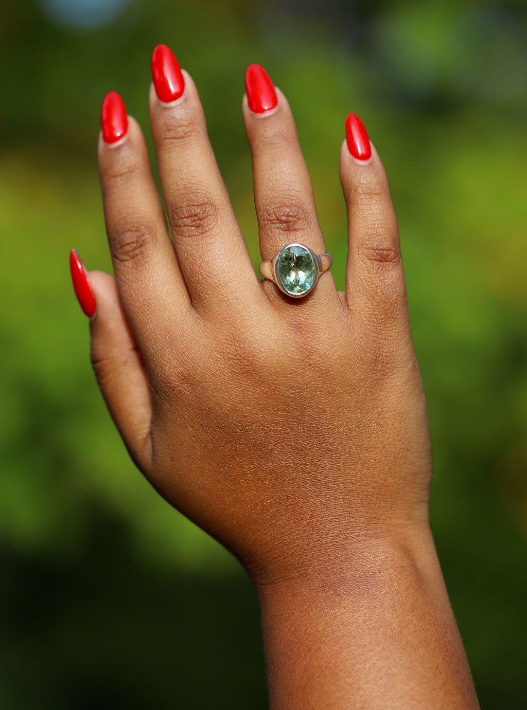 Hand with red nail polish wearing a green gemstone ring against a blurred green background