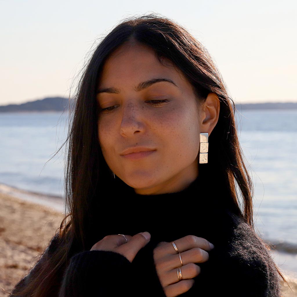 brunette looking down on beach wearing sterling silver three square post earrings and sterling silver wavy and flat rings