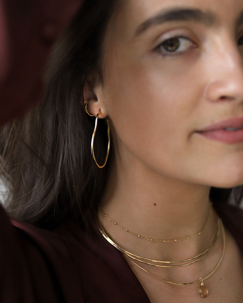 brunette wearing brass maeve thin hoop earrings champagne quartz gemstonce arc necklace and two 14k gold filled halo collars