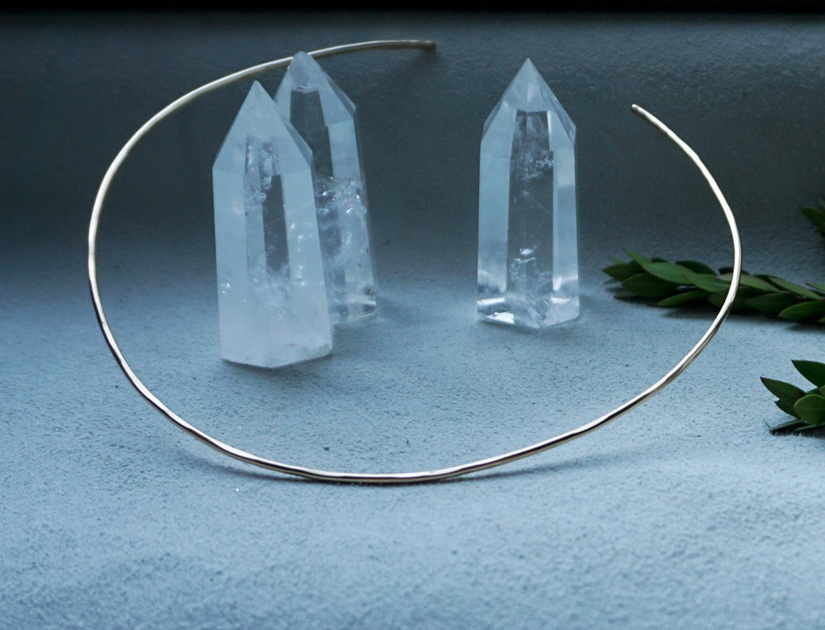 14k gold filled halo collar reflecting light on a grey surface surrounded by quartz crystals and a branch of green leaves