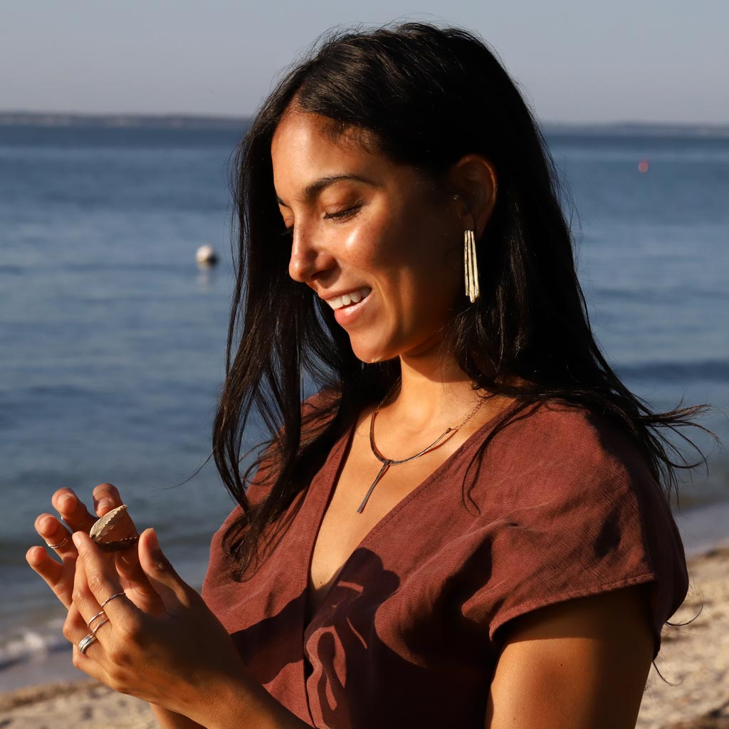 brunette at beach looking at shell on her hands wearing gold brass long fringe post earrings under sunlight