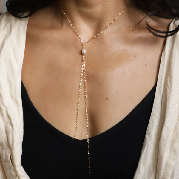 neck and chest with gold and pearl y shaped chain necklace against a dark background