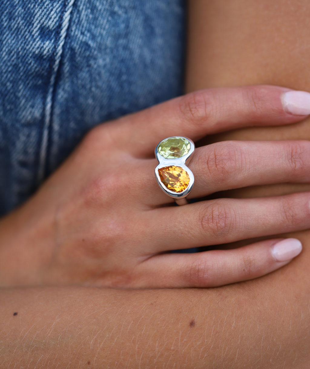 A close-up of a person's hand wearing a silver ring with two oval-cut gemstones, one light green and one amber-orange, against blue denim and skin tones.
