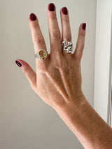Hand wearing two rings with a yellow gemstone and silver design against a neutral background