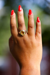 Hand with red nail polish wearing a ring with a yellow gemstone against a blurred natural background