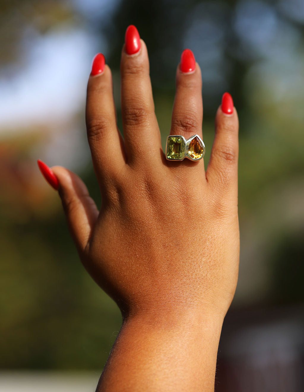 Hand with red nail polish wearing a green gemstone ring against a blurred natural background
