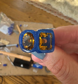Hand holding a blue ring with yellow gemstones on a wooden surface.