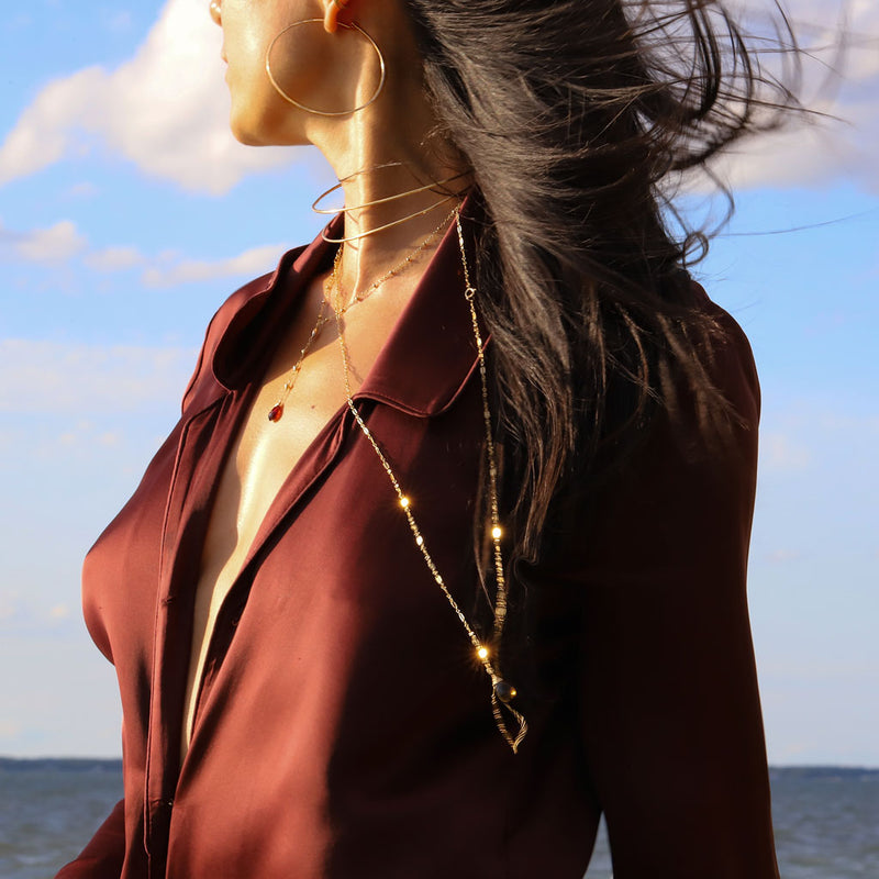Brunette woman wearing gold wrap necklaces with garnet gemstones and a red shirt on the beach 