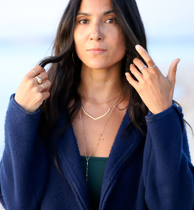 Brunette woman wearing gold layered necklaces, silver thin and big rings, and a blue sweater.
