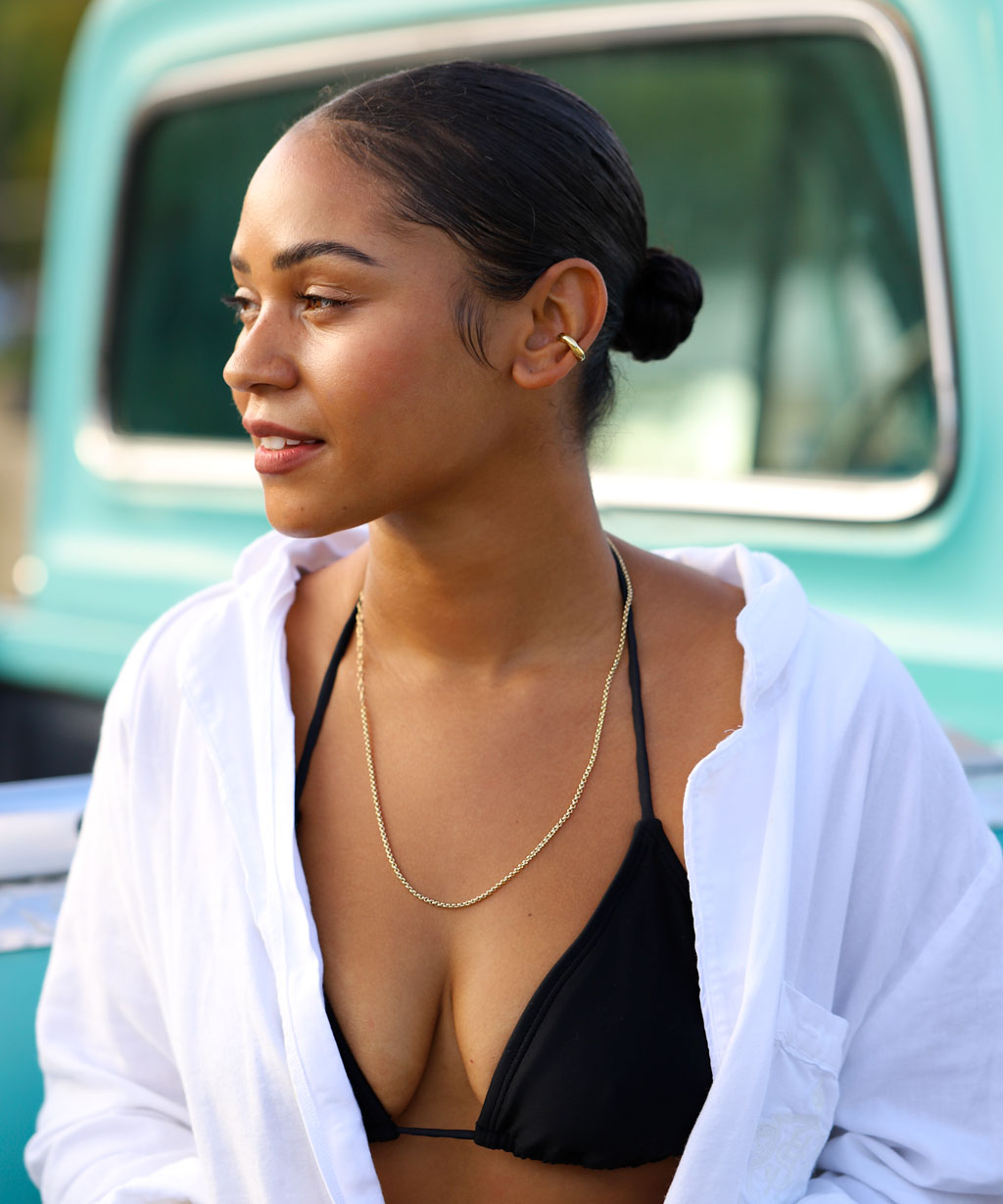 girl in black bikini top and loose white open shirt wearing a simple gold chain necklace and a gold cartilage hoop earring against a turquoise background