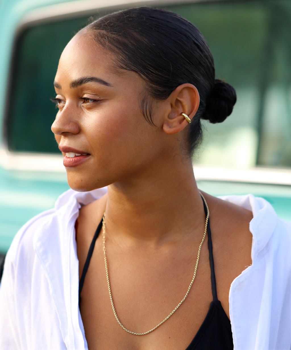 girl in black bikini top and loose white open shirt wearing a simple gold chain necklace and a gold cartilage hoop earring against a turquoise background