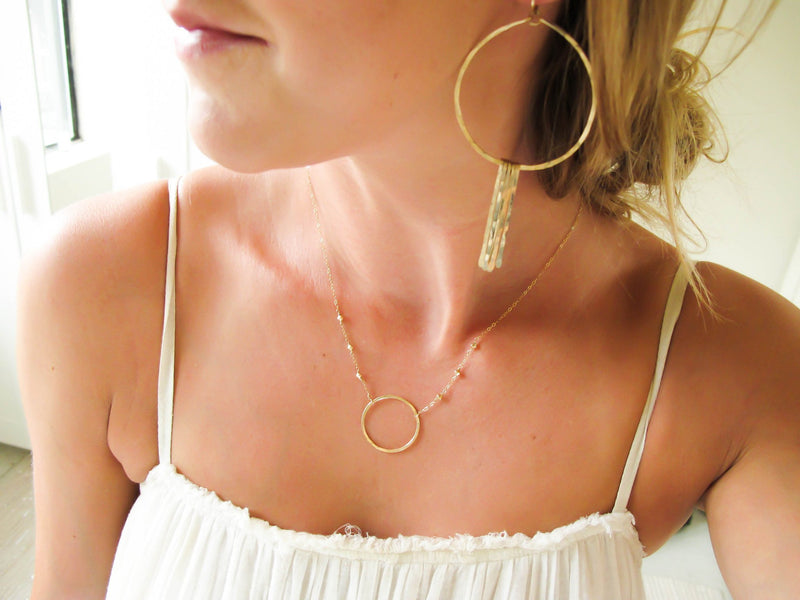 blond woman on a white top wearing a 14k gold filled xl unity beaded necklace with a gold circle pendant at home