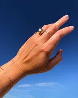 woman hand wearing globe ring on ring finger and thin gold stacking rings on middle and pinky fingers against blue sky