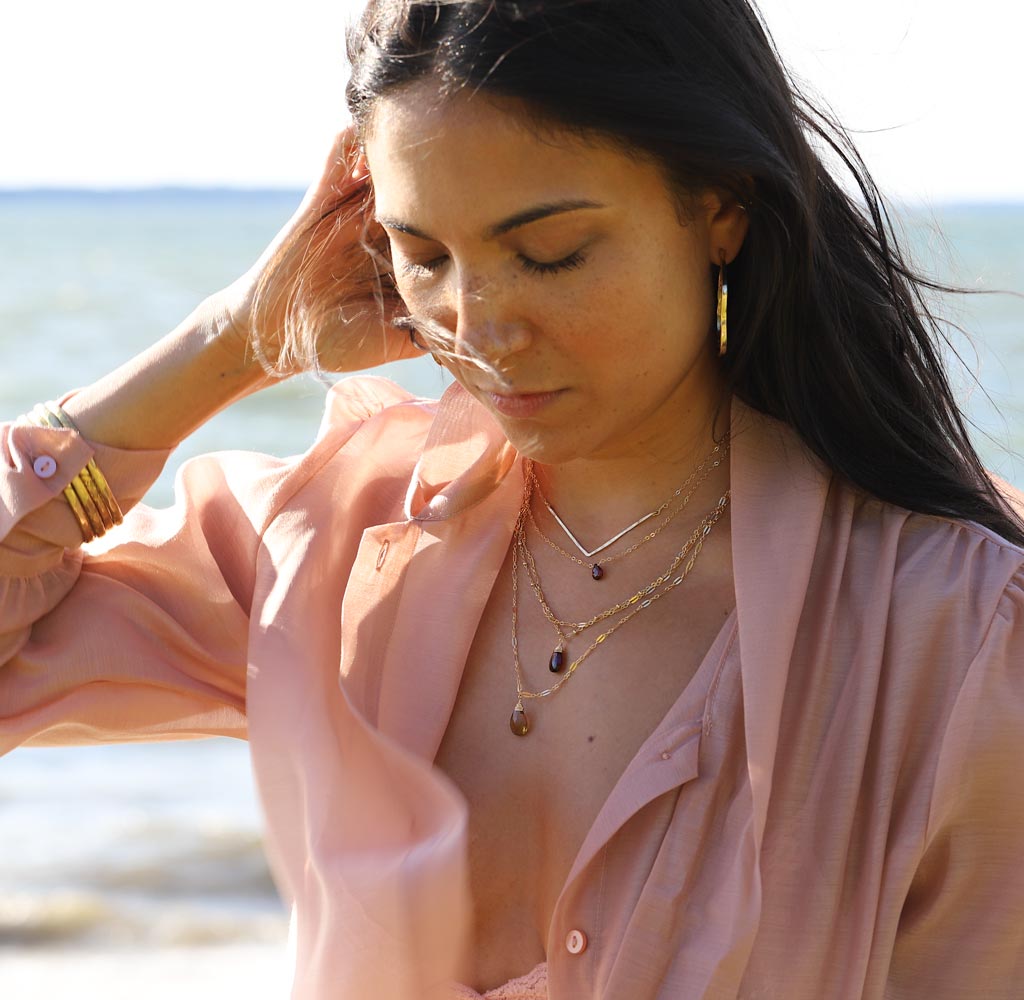 brunette at beach on a pink shirt against wind looking down wearing a 14k gold fill garnet short gemstone necklace