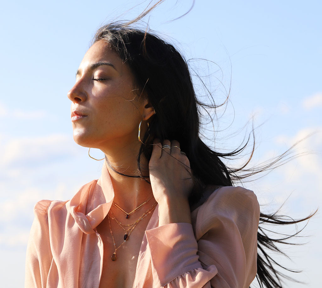 brunette at beach on a pink shirt against wind with eyes closed wearing a 14k gold fill garnet short gemstone necklace