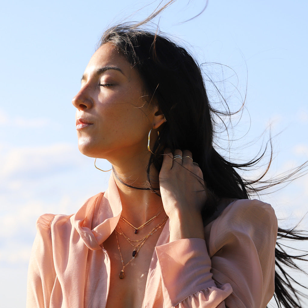 pink blouse brunette against wind wearing a garnet short gemstone necklace and garnet and beer quartz y gemstone necklaces
