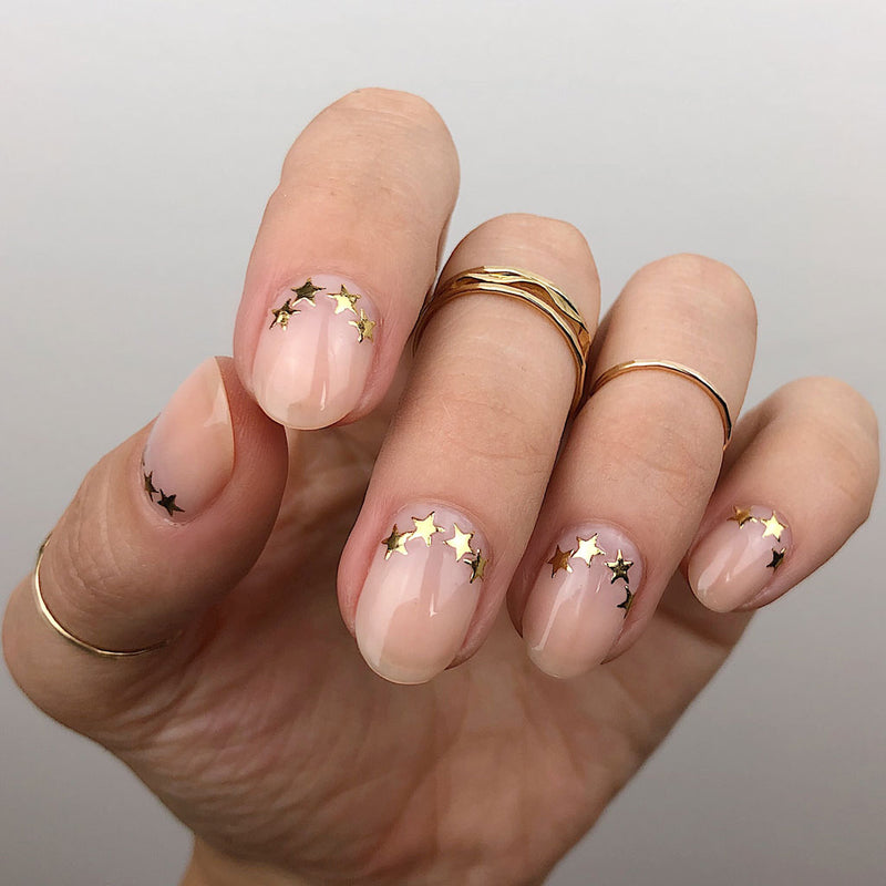 pink gold stars nails hand wearing 14k gold filled wavy and flat rings on thumb middle and ring fingers on a grey background