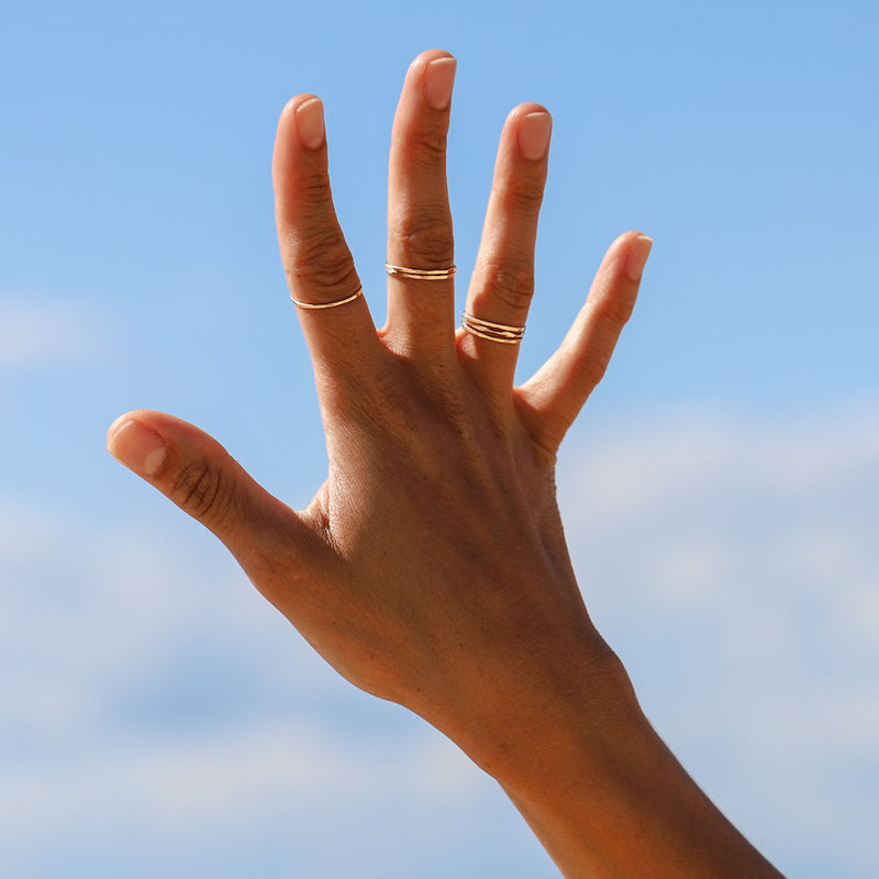 woman hand up in the air wearing 14k gold fill wavy and flat rings on index middle and ring fingers on a blue sky background