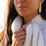 brunette holding her white wool sweater wearing 14k gold filled wavy and flat rings on both hands under sunlight at the beach