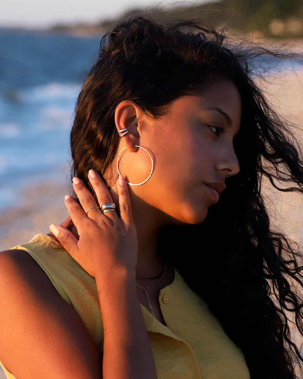 Brunette woman wearing silver hoops, hug hoops and big rings at the beach 