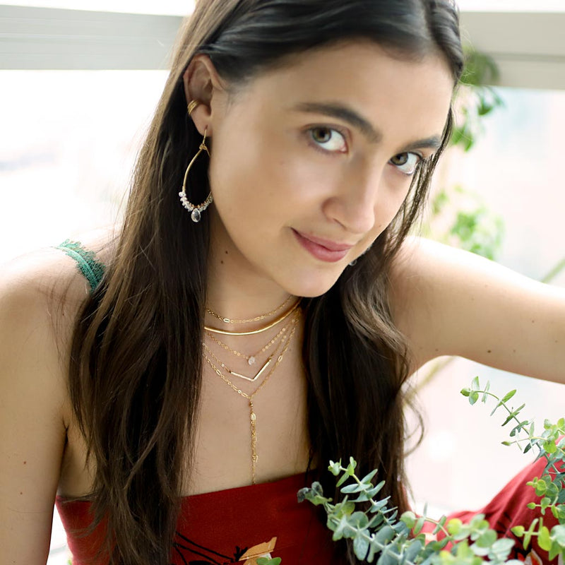 brunette surrounded by branches of leaves on a red top looking at camera wearing a 14k gold filled wide v necklace