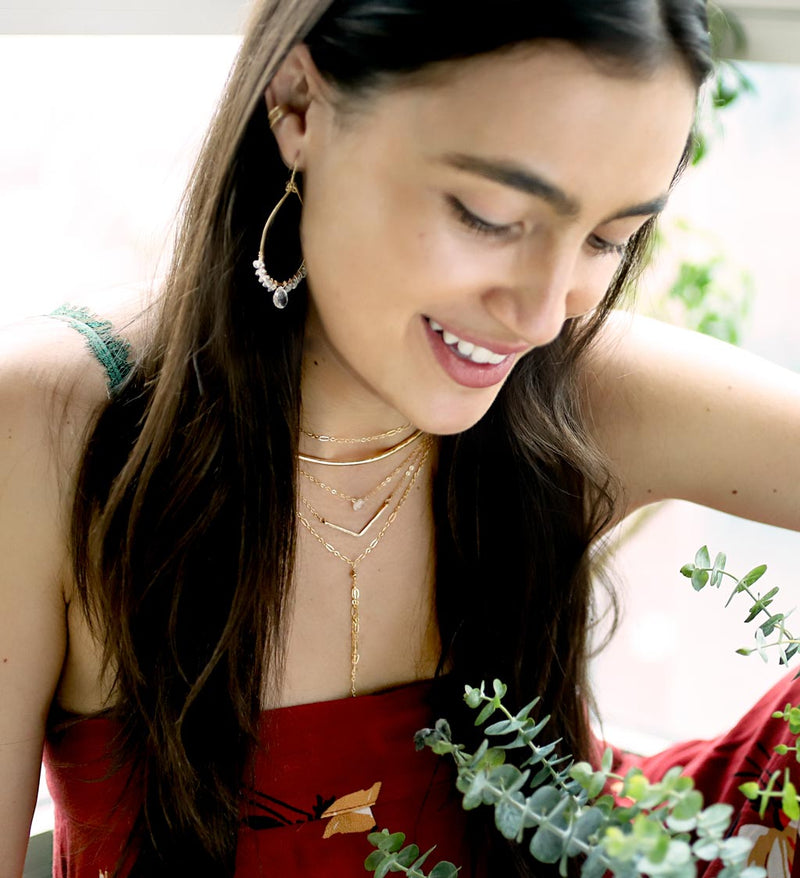 brunette surrounded by branches of leaves on a red top looking down wearing a 14k gold filled wide v necklace